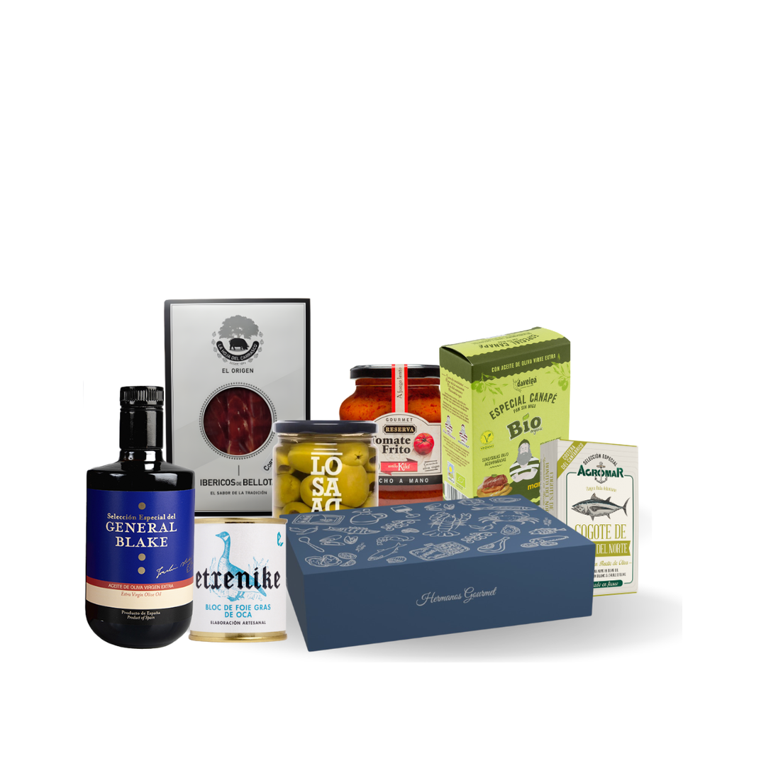Contenido de la caja regalo Grand Gourmet: jamón ibérico, foie gras de oca, aceite de oliva General Blake, cogote de bonito, tomate frito, tostas para canapés y aceitunas Manzanilla junto a la caja azul oscuro de Hermanos Gourmet