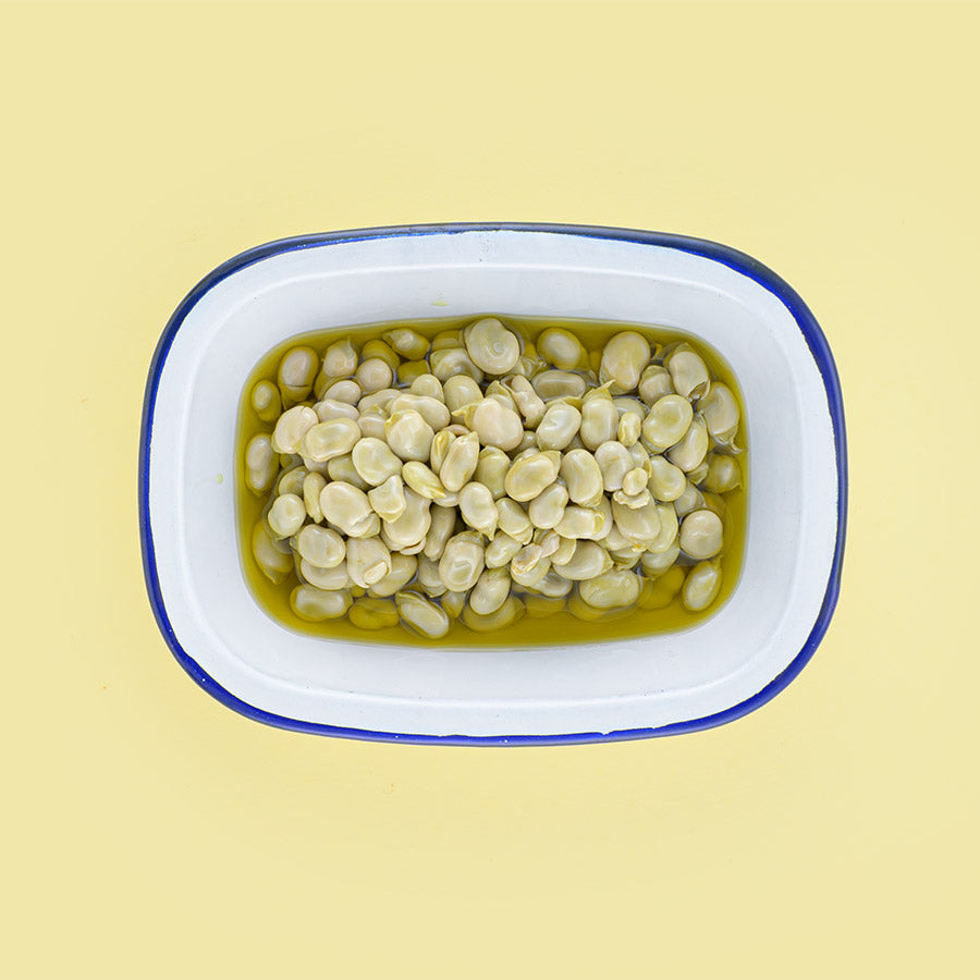 Petites fèves à l'huile d'olive dans un plat en émail blanc à bord bleu, sur fond jaune pâle